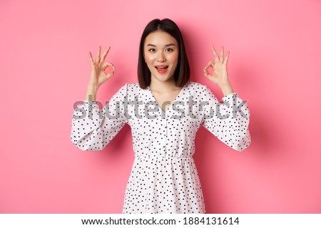 Pretty young asian woman in dress showing okay sign, praising and showing approval, looking satisfied, standing against pink background