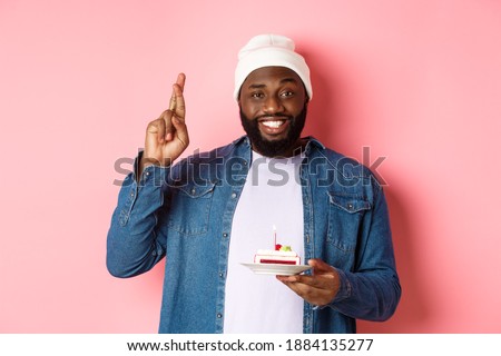 Handsome african-american guy celebrating birthday, making wish with fingers crossed, holding bday cake with candle, standing against pink background