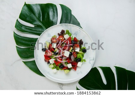 Close-up Traditional Greek salad with tropical monstera leaves under plate. Salad with Feta cheese, black olives, lettuce, fresh cucumber, cherry tomatoes served with red onion on marble background