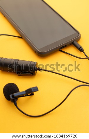 Lavalier or lapel microphone on a colored surface plugged into a cell phone. Details of the grip clip or fastener, mic power bank, miniplug, and wind sponge are visible. Concept sound production,