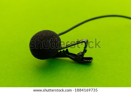 Lavalier or lapel microphone on a green surface, very close-up. The details of the grip clip or bra and the sponge against the wind are visible.