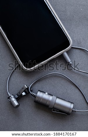 Lavalier or lapel microphone on a black surface, very close-up. The details of the grip clip or bra, , powerbank and conection to cellphone.