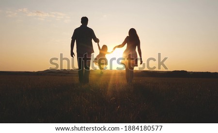 Child plays with dad and mom on field in sunset light. Family and childhood. Little daughter jumping holding hands of dad and mom in park on background of sun. Walking with a small kid in nature.