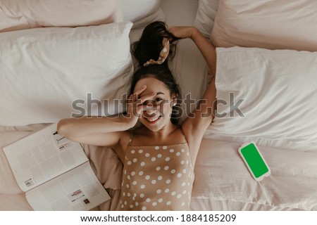 Brown-eyed brunette Asian woman in beige polka dot dress touches face, smiles and lies in bed. Cool lady poses near magazine and cellphone with green screen.