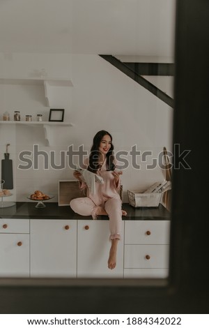 Cool brunette curly Asian woman with red lips dressed in pink trendy pajamas sits on kitchen table, smiles and holds newspaper.