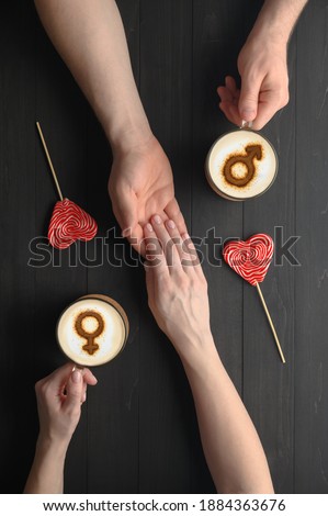 Two cups of coffee with symbols of venus and mars on milk foam and holding hands of couple in love on dark wooden table. Concept romantic date on Valentine's day. Top view, creative flat lay