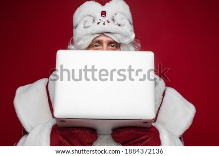 Santa Claus hiding behind laptop with copy space on red studio background, peek out checking xmas email accepting orders