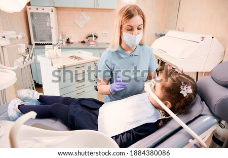 Young woman dentist in medical face mask holding dental instruments while little girl lying in dental chair with inhalation sedation. Concept of sedation dentistry. Royalty-Free Stock Photo #1884380086