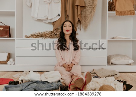 Pretty cool girl in pink pajamas looks up dreamily. Attractive woman with red lips sits in dressing room with plenty of clothes on wooden floor.