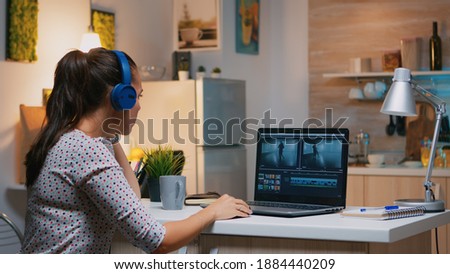 Freelancer content creator working overtime from home to respect deadline. Woman videographer editing audio film montage on professional laptop sitting on desk in modern kitchen in midnight