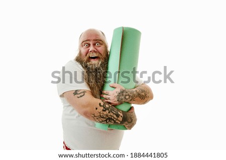 A bearded overweight man with tattooed arms gently hugs his fitness mat and laughs.