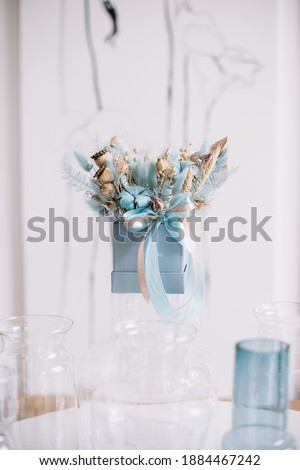 Beautiful gift composition an a beautiful blue box with dry flowers in it, standing on a glass vase on the white wall background 