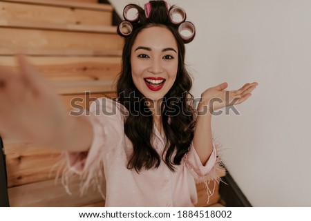 Excited cool brown-eyed woman with hair curlers and red lips takes selfie. Pretty girl in pink pajamas smiles sincerely at home.