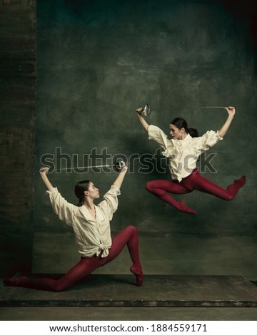 Flying. Two young female ballet dancers like duelists with swords on dark green background. Caucasian models dancing together. Ballet and contemporary choreography concept. Creative art photo.