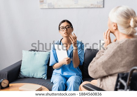 asian nurse with digital tablet and elderly woman touching faces while talking in hospital, blurred foreground