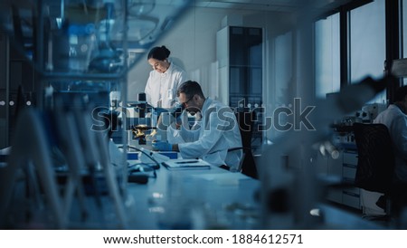 Modern Medical Research Laboratory: Two Scientists Working, Using Digital Tablet, Analyzing Test, Talking. Advanced Scientific Pharmaceutical Lab for Medicine, Biotechnology Development. Evening Time Royalty-Free Stock Photo #1884612571