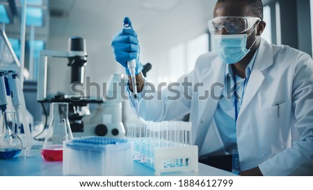 Medical Development Laboratory: Black Scientist wearing Face Mask Uses Pipette for Filling Test Tube with Liquid, Conducting Experiment. Pharmaceutical Lab with Medicine, Biotechnology Researchers Royalty-Free Stock Photo #1884612799