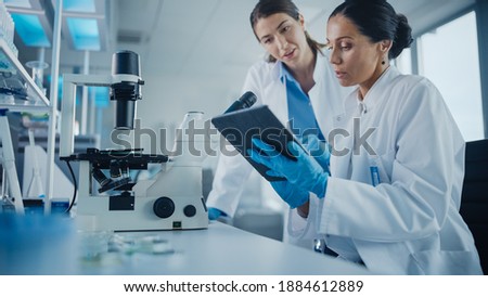 Modern Medical Research Laboratory: Two Female Scientists Working, Using Digital Tablet, Analysing Samples, Talking. Advanced Scientific Pharmaceutical Lab for Medicine, Biotechnology Development Royalty-Free Stock Photo #1884612889