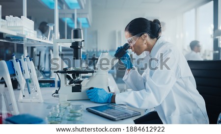 Medical Development Laboratory: Caucasian Female Scientist Looking Under Microscope, Analyzes Petri Dish Sample. Specialists Working on Medicine, Biotechnology Research in Advanced Pharma Lab Royalty-Free Stock Photo #1884612922