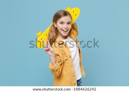 Funny little blonde kid girl 12-13 years old in casual yellow jacket posing isolated on blue background studio. Childhood lifestyle concept. Mock up copy space. Hold skateboard, showing victory sign