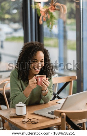 joyful african american woman holding cup while looking at laptop in cafe