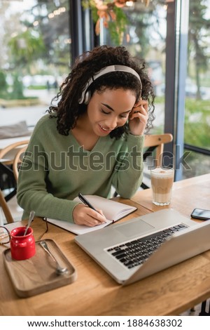 happy african american woman listening podcast in headphones and writing in notebook near laptop in cafe Royalty-Free Stock Photo #1884638302