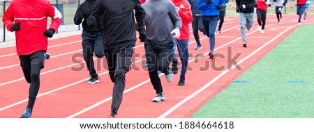 A High school boys winter track team running on a track covered up with spandex and sweatpants on their legs, sweatshirts and gloves to help keep them warm. Royalty-Free Stock Photo #1884664618