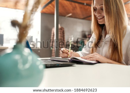 Creative female. A cute middle age blonde fashionably dressed smiling and sitting at a table making notes in her flipbook with a pen in front of a laptop in a bright fashion workshop