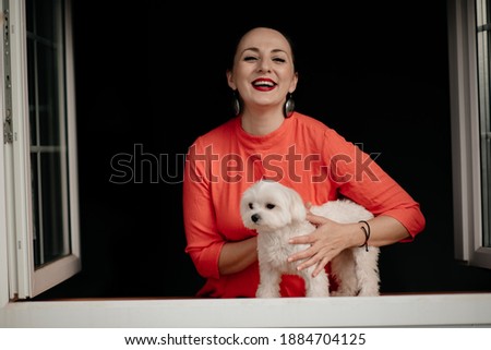 Happy smiling girl with black hair in a red dress with stylish earrings in her ears polishes with her little white dog on a black background.
