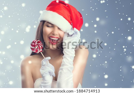 Christmas woman. Beauty model girl in Santa Claus hat with red lips and xmas lollipop candy in her hand. Joy. Surprised expression. Closeup portrait over winter snow background 
