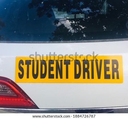 A yellow student driver sticker on a white car. The rear windshield and taillight is also seen.