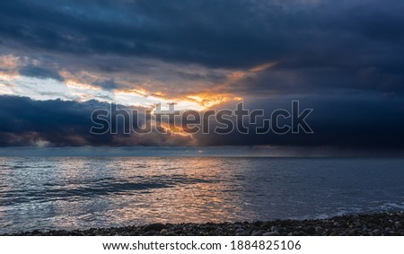 Alarming stormy sunset over the sea. Dense dark clouds cover the sky. Their gap is illuminated by orange rays of the sun. It's raining in the distance. Reflection on a shiny water surface. Black Sea