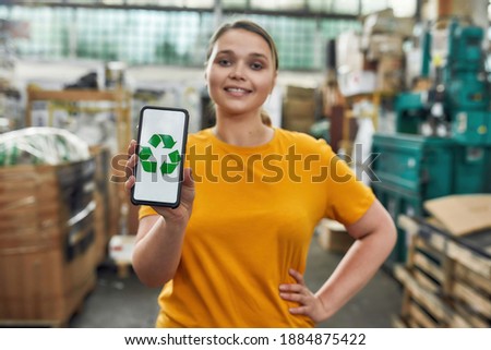 Selective focus on smartphone screen showing recycling sign with green arrows in hand of smiling caucasian young girl working on garbage station. Waste sorting and recycling concept