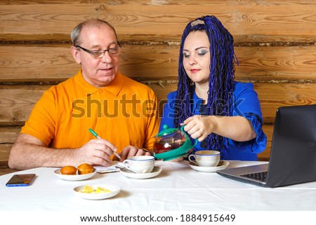 A family couple a man in glasses and a woman with blue afro braids at the table a woman pours tea a man writes information in a notebook. Horizontal photo