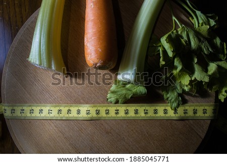 Overhead view photo of a wooden cutting board with a carrot, celery and a measuring tape on it, symbolizing a healthy and balanced diet.