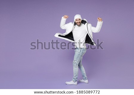 Full length side view of funny man in white padded windbreaker jacket hat turn around spreading rising hands dancing isolated on purple background studio. People lifestyle cold winter season concept