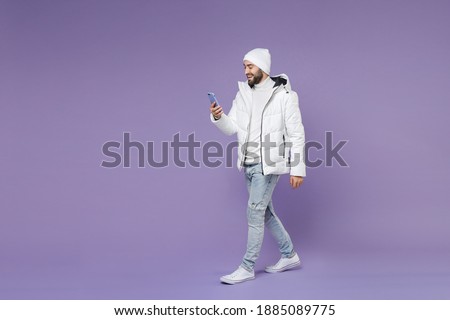 Full length side view smiling man in warm white windbreaker jacket hat using mobile cell phone typing sms message isolated on purple background studio portrait. People lifestyle cold season concept