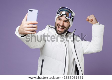 Happy skier man in white jacket ski goggles mask doing selfie shot on mobile phone doing winner gesture spend weekend winter in mountains isolated on purple background. People lifestyle hobby concept