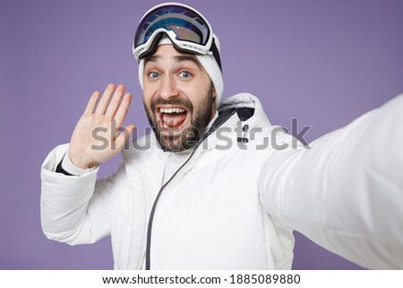 Close up of amazed skier man in windbreaker jacket ski goggles mask doing selfie shot on mobile phone greeting with hand spend weekend in mountains isolated on purple background. People hobby concept