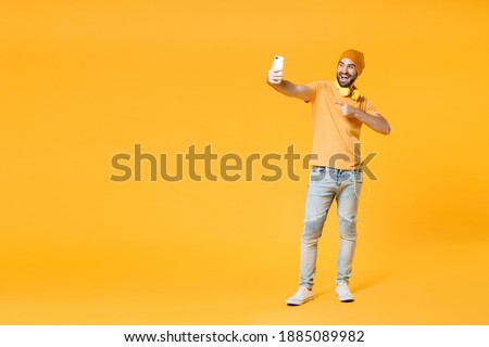 Full length of funny young bearded man 20s wearing basic casual t-shirt headphones hat doing selfie shot pointing index finger on mobile phone isolated on bright yellow background, studio portrait