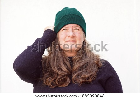 Middle aged woman wearing black sweater and green beanie on a chilly day outside against a white background holds her hat with one hand while looking up and to the right                              