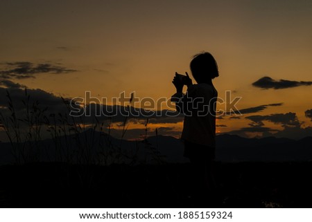 Silhouette of child girl holding a smartphone taking pictures at meadow on sunset time.