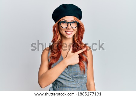 Young redhead woman wearing fashion french look with beret cheerful with a smile on face pointing with hand and finger up to the side with happy and natural expression 