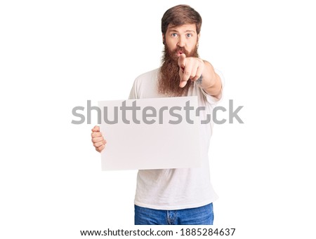 Handsome young red head man with long beard holding blank empty banner pointing with finger to the camera and to you, confident gesture looking serious 