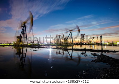 Oil field site, in the evening, oil pumps are running, The oil pump and the beautiful sunset reflected in the water, the silhouette of the beam pumping unit in the evening.