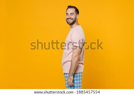 Side view of smiling young bearded man 20s in pajamas home wear looking camera while resting at home isolated on bright yellow colour background studio portrait. Relax good mood lifestyle concept