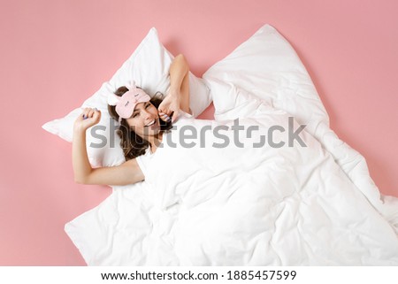 Overhead top view of funny young woman in pajamas home wear sleep mask lying stretching hands resting at home isolated on pastel pink background studio portrait. Relax good mood lifestyle concept