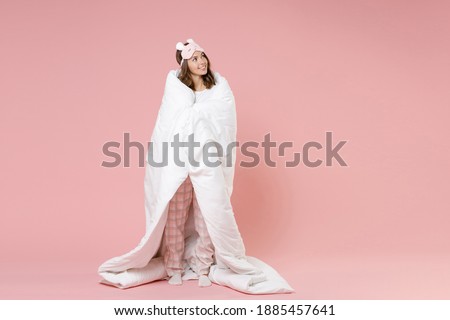 Full length portrait of smiling pretty cute young woman in pajamas home wear sleep mask wrapping in blanket looking aside resting at home isolated on pink background studio . Relax good mood concept