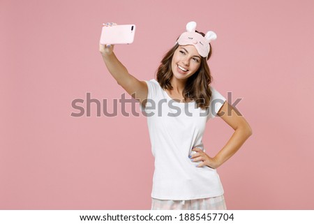Cheerful young brunette woman wearing white pajamas home wear sleep mask doing selfie shot on mobile phone resting at home isolated on pastel pink background studio portrait. Relax good mood concept
