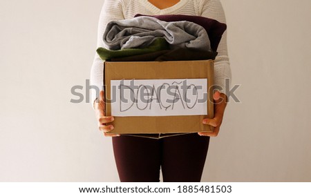 Donation written in Portuguese (Doação) on a cardboard box. Woman holding a Donation Box full of clothes.